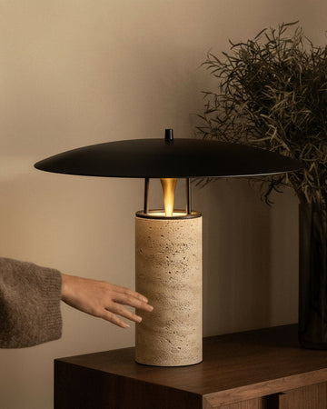 Person adjusting a modern table lamp on a wooden side table with a vase of dried plants.